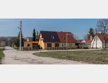 Ein gelbes Einfamilienhaus mit rotem Satteldach steht an einer Kreuzung in einer ländlichen Umgebung. Im Vordergrund befinden sich eine unbefestigte Straße, ein Straßenschild und eine Rasenfläche, im Hintergrund sind weitere Häuser und Baumreihen zu sehen.