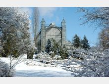 Ein historisches Gewächshaus mit hohen, gläsernen Türmen, umgeben von verschneiten Bäumen und Sträuchern in einer winterlichen Gartenlandschaft unter klarem blauem Himmel.