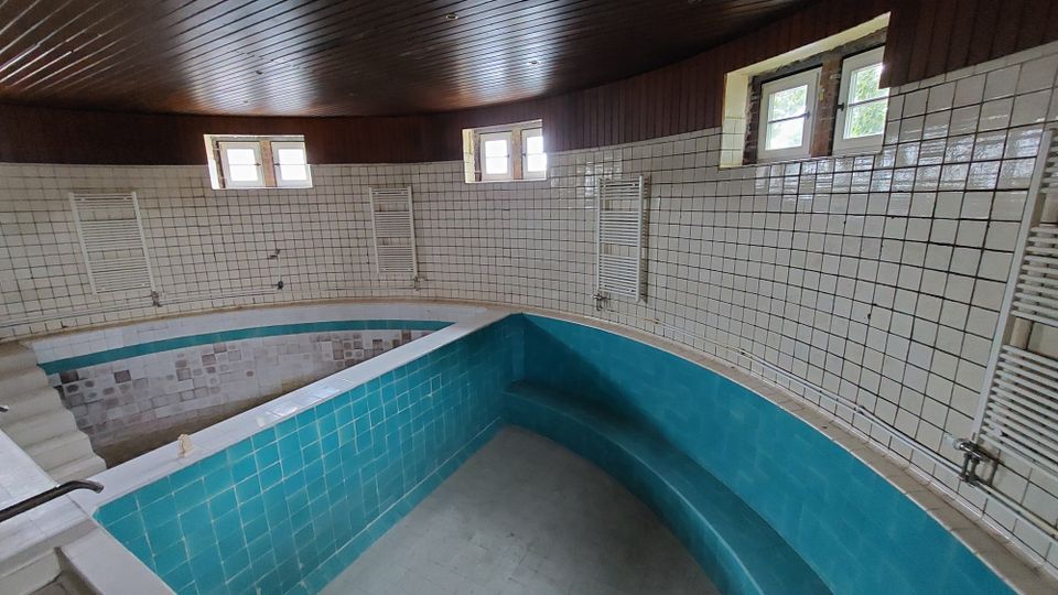 Innenansicht eines leeren, gefliesten Pools mit türkisfarbenen Kacheln und umliegenden weißen Wänden, die mit kleinen Quadratkacheln verkleidet sind. Oben sind kleine Fenster und eine Holzdecke sichtbar.