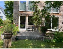 Holzhaus mit großer Terrasse, umgeben von Gartenpflanzen. Die Terrasse ist mit Blumenkübeln, Kletterpflanzen und einem kleinen Tisch mit Stühlen dekoriert. Im Hintergrund sind große Fenster und eine Balkontür sichtbar.