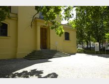 Gelbes Gebäude mit rechteckigen Fenstern und massiver Holztür, erreichbar über eine Treppe mit Metallgeländer, umgeben von einem gepflasterten Vorplatz und schattenspendenden Bäumen. Ein Informationsschild steht rechts vor dem Gebäude.