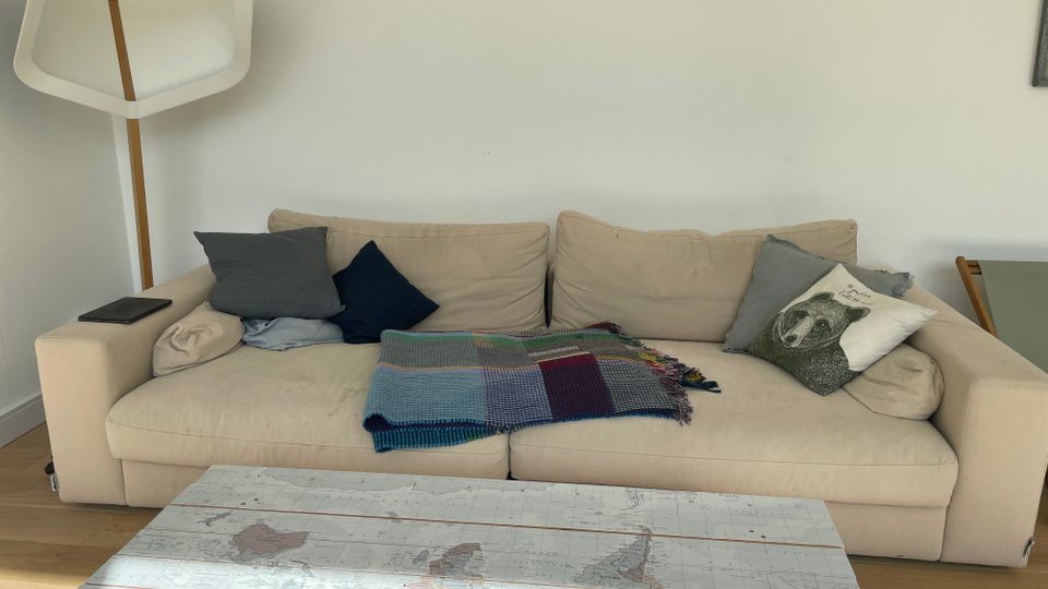 Wohnzimmer mit beigem Sofa, mehreren Kissen und einer karierten Decke; links steht eine weiße Stehlampe mit Holzstiel. Im Vordergrund liegt ein Couchtisch mit Weltkarten-Motiv, dahinter eine weiße Wand mit schmaler Ablage und Regalfach darüber.