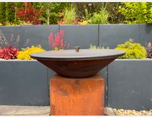 Eine moderne Feuerschale aus Metall auf einem rostfarbenen Sockel, umgeben von einer terrassierten Gartenlandschaft mit grauen Wänden, bunten Blumen und grünem Gebüsch.