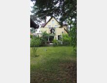 Ein gelbes Einfamilienhaus mit Satteldach, umgeben von einem Garten mit Rasenfläche und Bäumen; an der Hausfassade hängt ein großes Poster mit der Aufschrift "One Love" und einem Bild von Bob Marley.