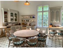 Rustikaler Küchenraum mit einem großen runden Holztisch und passenden Holzstühlen mit gepolsterten Sitzflächen. Die Wände sind mit offenen Regalen, weißen Schränken mit Gitterfronten und großen Fenstern ausgestattet, die viel Tageslicht hereinlassen; der Boden hat ein gemustertes Fliesendesign.