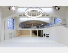 Heller, moderner Innenraum mit einer hohen Decke, großen Bogenfenstern und zwei Ebenen, wobei die obere Ebene über eine gläserne Brüstung verfügt. Ein großer, runder Kronleuchter dominiert die Decke, während die Wände mit eingelassenen Regalen und dezenten Vorhängen gestaltet sind.