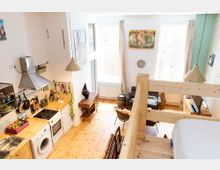 Heller Innenraum einer kleinen Wohnung mit Holzfußboden und einer offenen Galerie. Im unteren Bereich befinden sich eine Küche mit Holzarbeitsflächen, ein Wohnbereich mit Ledersessel und dekorativen Elementen sowie große Fenster, die Tageslicht hereinlassen.