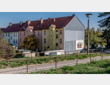 Mehrfamilienhäuser mit farbigen Fassaden und einem angebauten Gebäude mit Wellblechverkleidung, auf dem eine Werbetafel angebracht ist. Im Vordergrund ein Fußweg mit Geländer und vegetationsbewachsene Grünflächen, im Hintergrund Bäume und ein Straßenbereich.