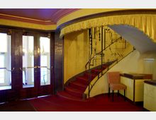 Innenbereich eines Theaters oder Kinos im Art-Déco-Stil mit gelb gestrichenen Wänden, rotem Teppichboden und einer geschwungenen Treppe mit filigranem Metallgeländer. Links sind Holztüren mit Glaseinsätzen zu sehen, rechts steht ein gepolsterter Stuhl neben einem Empfangstresen.