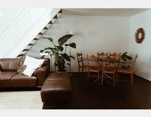 Wohnzimmer mit hellem, minimalistischem Design: braunes Ledersofa mit Kissen links, daneben eine große Grünpflanze. Rechts befindet sich ein Esstisch aus Glas mit Holzstühlen. Eine weiße Treppe führt nach oben, und an der Wand hängt ein runder Dekorationsspiegel.