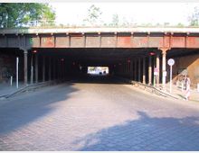 Eine gepflasterte Straße führt unter einer alten Brücke hindurch, die von Metallträgern und Säulen gestützt wird. Auf beiden Seiten der Unterführung sind rote Warnlichter und Verkehrsschilder zu sehen, umgeben von Graffiti an den Wänden.