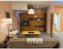 Wohnzimmer im Retro-Stil mit orange-brauner Tapete im 70er-Jahre-Muster, Holzschrankwand mit Fernseher und Stereoanlage, gemustertem Sofa, Couchtisch und Stehlampe mit besticktem Lampenschirm. Im Hintergrund führt eine Tür zu einem angrenzenden Raum.