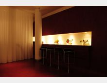 Modernes Innenraumdesign mit einer langen Theke, die durch dezente integrierte Beleuchtung hervorgehoben wird. Vor der Theke befinden sich vier schwarze Hocker, im Hintergrund hängen weiße Vorhänge und eine runde Säule gliedert den Raum.