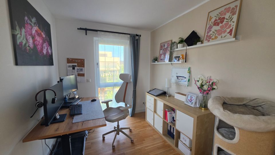 Helles, schmales Homeoffice mit Holzfußboden, Schreibtisch mit zwei Monitoren links und Regal-/Schrankkombination mit Deko und Pflanzen rechts. Am Ende des Raums sorgt eine bodentiefe Glastür mit Vorhängen für Tageslicht; an den Wänden hängen Blumenbilder und ein Pinboard.