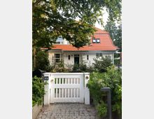 Zweistöckiges Einfamilienhaus mit weißer Fassade und rotem Satteldach, umgeben von einem gepflegten Garten. Im Vordergrund ein weißes Holztor mit Weg aus Kopfsteinpflaster, Bäume und Büsche rahmen die Szene ein.