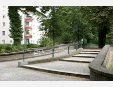 Pflasterweg mit Stufen und angrenzender Rampe mit grünem Geländer, umgeben von Bäumen und Hecken. Im Hintergrund befindet sich ein mehrstöckiges Wohnhaus mit Balkonen.