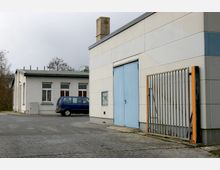 Industriegebäude mit heller Fassade, einem großen blauen Tor und angrenzendem Metallzaun. Im Hintergrund steht ein kleineres Flachdachgebäude mit mehreren Fenstern und einem parkenden blauen Fahrzeug.