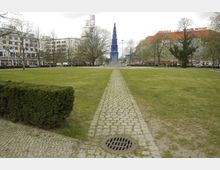 Grüne Parkanlage mit einem gepflasterten Weg, der auf einen hohen, blauen, stufenförmigen Obelisken führt. Im Hintergrund sind mehrstöckige Gebäude, darunter ein weißes Geschäftshaus und ein rotdachiges Gebäude, sowie vereinzelte Bäume sichtbar.