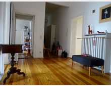 Heller Wohnungsflur mit Holzdielenboden, weißen Wänden und mehreren Türen, die zu angrenzenden Räumen führen. Ein kleines, antikes Holztischchen, ein graues Möbelstück mit dekorativen Gegenständen und ein gelber Stuhl ergänzen die Einrichtung.