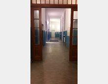 Ein langer, geradliniger Innenflur in einem Gebäude mit hellen Wänden und Holzrahmen-Doppeltüren im Vordergrund. Die Wände des Flurs sind in der unteren Hälfte blau gestrichen, und der Boden ist aus dunklem Linoleum; helle Lampen beleuchten den Gang, in dem sich mehrere Türen und ein Fenster befinden.