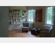 Ein gemütlicher Leseraum mit Bücherregalen an der Wand, zwei gepolsterten Sesseln, einem kleinen Couchtisch mit Büchern und einer Stehlampe. Zwei große Fenster lassen Tageslicht in den Raum und bieten einen Blick ins Grüne.