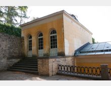 Gelbes, historisches Gebäude mit drei hohen, bogenförmigen Fenstern und verzierten weißen Türen, flankiert von einer Steinmauer und einer Treppe. Im Vordergrund befindet sich ein Geländer aus Metall, daneben ist ein angebautes Dach mit Glas- und Metalldetails sichtbar.