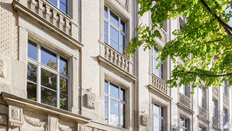 Mehrstöckiges, historisches Stadthaus mit heller Backsteinfassade und rustiziertem Sockelgeschoss, großen rechteckigen Fenstern und einem Rundbogen-Eingang. Im Vordergrund verläuft ein Gehweg entlang der Straße, rechts ragen grüne Baumkronen ins Bild.