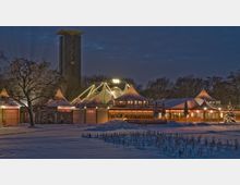 Winterliche Abendaufnahme eines beleuchteten Veranstaltungsortes mit weißen Zeltkonstruktionen, darunter das Tipi-Zelt. Im Hintergrund ragt ein moderner Glockenturm empor, umgeben von kahlen Bäumen und schneebedecktem Gelände.