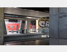 Bahnhof Warschauer Straße in einer urbanen Umgebung, mit sichtbaren Betonpfeilern und Graffitis auf den Wänden. Im Hintergrund fährt ein roter Regionalzug vorbei, während Fahrräder und Abfallbehälter am Bahnsteig stehen.