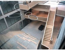 Innenansicht eines modernen Gebäudes mit offener Mehrzweckhalle. Sichtbar sind Treppen, hölzerne Sitzstufen, eine Tischtennisplatte auf Betonboden, umlaufende Glastüren sowie hängende Ringlampen als Beleuchtung.