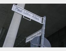 Straßenschilder zeigen die "Paul-Löbe-Allee" und die "Konrad-Adenauer-Straße" an einer Kreuzung. Im Hintergrund sind Betonpfeiler und eine moderne Glasfassade eines Gebäudes zu sehen.