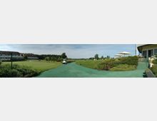 Panoramaaufnahme einer gepflegten Golfanlage mit einem angrenzenden Hotelgebäude links im Hintergrund, Rasenflächen und Büschen sowie einem Golfplatzweg in der Mitte. Rechts im Vordergrund befinden sich Stufen, ein Sonnenschirm und bepflanzte Beete.