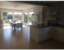 Moderne Wohnküche mit offenem Essbereich, großen bodentiefen Fenstern und Blick in einen grünen Garten. Die Küche verfügt über helle Fronten, ein dunkles Arbeitsplatten-Design und Wandregale mit dekorativen Elementen.