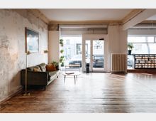 Heller, minimalistischer Innenraum mit Holzboden, einer grünen Couch an einer teils abgenutzten Wand, und Glas-Doppeltüren, die nach außen führen. Pflanzen, Lampen und ein kleiner Tisch ergänzen die Einrichtung; draußen sind Autos sichtbar.