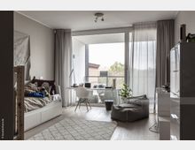 Ein modernes, helles Zimmer mit einem Tagesbett links, einem Schreibtisch und Stuhl vor einer großen Glas-Schiebetür, die auf einen Balkon mit Blick auf Bäume und ein Gebäude führt. Der Raum ist mit hellen Vorhängen, einem Teppich und einer Sitzsack-ähnlichen Sitzgelegenheit ausgestattet.