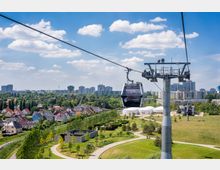 Seilbahn über einem parkähnlichen Gelände mit Rasenflächen, Bäumen und geschwungenen Gehwegen; im Hintergrund eine Stadtsilhouette mit Wohnhäusern und Hochhäusern sowie blauem Himmel mit einigen Wolken.