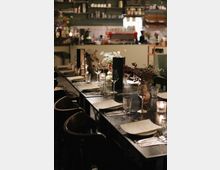 Ein gemütlich beleuchteter Bistro-Innenraum mit gedecktem Tisch, auf dem Gläser, Menükarten und Kerzen arrangiert sind; im Hintergrund eine offene Bar mit Regalen, Gläsern und einer Kaffeemaschine.