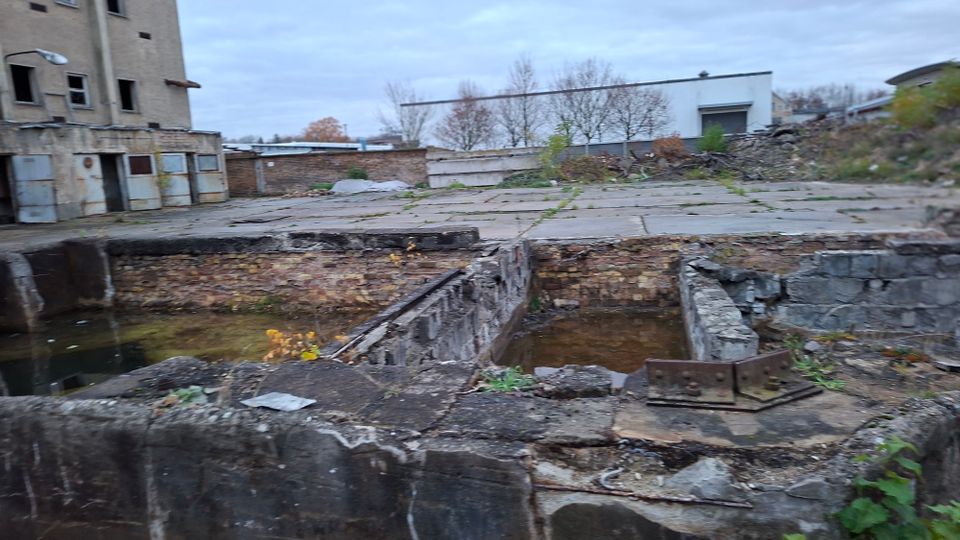 Verlassenes Industriegelände mit einer alten, teils eingestürzten Backsteinstruktur und einem offenen, mit Wasser gefüllten Graben; im Hintergrund stehen ein leerstehendes Gebäude und weitere Lagerhallen mit Bäumen und bewachsenem Boden.