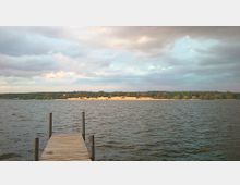 Holzsteg, der in einen ruhigen See führt, im Vordergrund. Im Hintergrund erstrecken sich Wälder und eine beleuchtete, goldfarbene Uferlinie unter einem teilweise bewölkten Himmel.