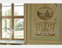 Großes Holzfenster mit Ausblick auf eine parkähnliche Landschaft, daneben eine verzierte Wandmalerei mit floralem Dekor und einer Landschaftsszene in einem ovalen Rahmen. Auf der Fensterbank steht ein silberner Kerzenständer mit fünf Armen.