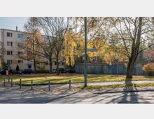 Wohngebiet mit mehrstöckigem Wohnhaus im Hintergrund, davor Bäume mit herbstlichem Laub. Rechts sind mehrere Garagen zu sehen, im Vordergrund eine Rasenfläche mit Gehweg und Straßenlaterne mit Straßenschildern.