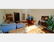 Wohnzimmer mit Holzboden, einer großen grauen Eckcouch und einem Holztisch im Vordergrund. An der Wand befinden sich ein Fernseher auf einer Retro-Kommode, ein Wandbild und Pflanzen; im Hintergrund steht ein alter Kachelofen neben einer Tür mit Glaseinsatz.