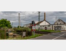 Bahnübergang mit Schranken und Andreaskreuz, daneben ein Industriegelände mit weißen Gebäuden, einer hohen Schornsteinsäule und Graffiti an den Wänden. Im Hintergrund verlaufen die Bahngleise mit Oberleitungen, umgeben von grüner Vegetation.
