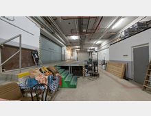 Industriehalle mit Betonboden, hohen Decken und sichtbaren Rohrleitungen an der Decke. Im Vordergrund befinden sich Stühle, Tische und eine kleine erhöhte Plattform mit Treppe; im Hintergrund sind Industrietore, eine Rampe und diverse Lagergegenstände zu sehen.