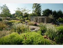 Naturgarten mit dichter Bepflanzung, verschiedenen Gräsern und Blüten sowie einem zentralen Steinplatz mit Feuerstelle. Im Hintergrund steht eine alte, verputzte Hütte mit Holztüren, umgeben von Bäumen und Sträuchern.