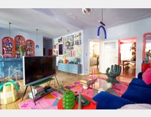 Eclectic, colorful living room with light wood floors, pale blue walls, and a large blue sofa around a glass coffee table on a bright patterned rug. The space features a gallery wall of framed art, arched shelving filled with bottles, modern pendant lights, and open double doors leading to a hallway and another room.