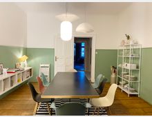 Esszimmer mit einem rechteckigen Tisch, umgeben von bunten Stühlen auf einem gestreiften Teppich. Der Raum hat Holzboden, halbe grün gestrichene Wände, eine weiße Tür, ein Wandregal mit Geschirr und niedrige Regale mit Büchern und Dekorationen. Drei Hängelampen leuchten den Raum aus.