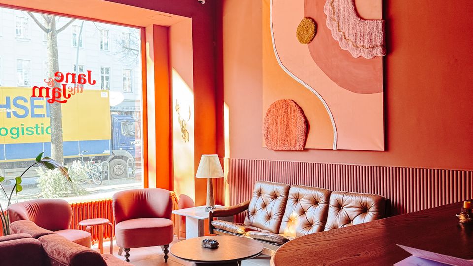 Cozy café lounge with warm pink and orange walls, a large street-facing window, and a seating area with upholstered sofas and armchairs around small round tables. Track lighting hangs from the ceiling, and an abstract wall artwork is mounted above a tufted bench seat next to a curved wooden bar counter.