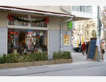 Straßenszene mit einem kleinen Restaurant oder Café an einer Hausecke. Die Fassade ist teils mit Wellblech verkleidet, und das Fenster zeigt bunte Neonreklamen sowie eine Dekoration mit Girlanden und roten Schleifen. Davor stehen bepflanzte Kästen, ein Werbeschild mit Menü-Angeboten und eine Gruppe von Fußgängern auf dem Gehweg.