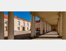 Neoclassical courtyard framed by a long covered colonnade with white columns and ornate capitals casting shadows on a stone walkway. Across the cobblestone square are pale stucco buildings with red tile roofs and tall arched windows under a clear blue sky.